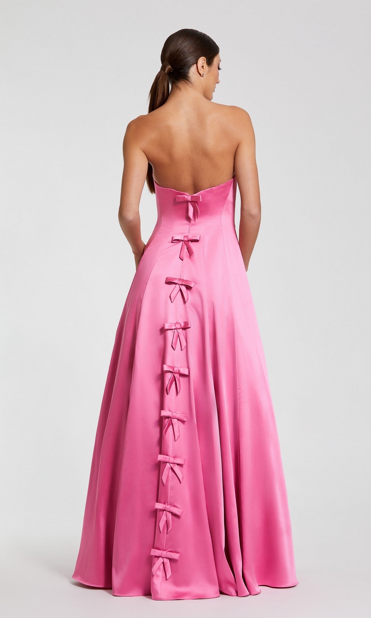 A woman poses against a plain light background wearing the Mac Duggal 68857 long formal dress, a strapless floor-length pink satin gown featuring multiple bows down the back.